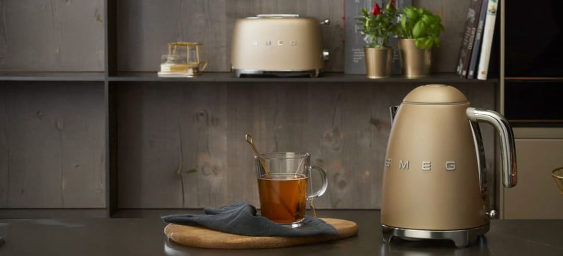 Bollitore elettrico Smeg color oro satinato accanto a una tazza di tè su un bancone da cucina moderno, con tostapane coordinato sullo sfondo.
