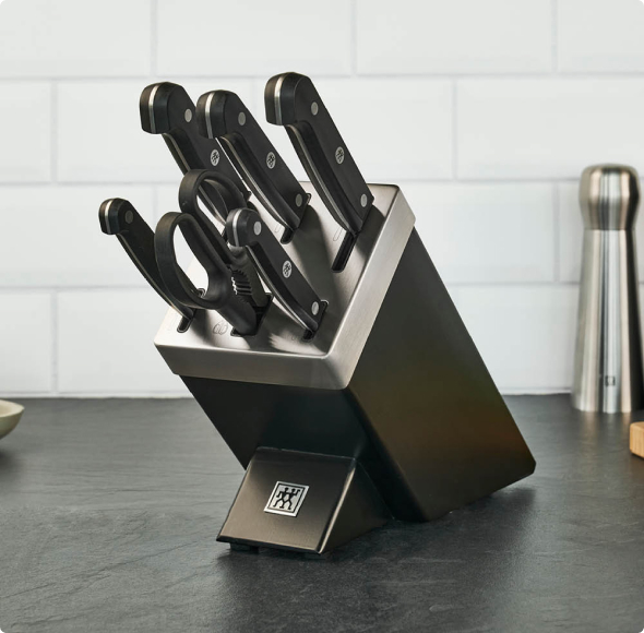 Set di coltelli professionali con lame affilate nel supporto moderno, utensili da cucina su piano in laminato e sfondo bianco.