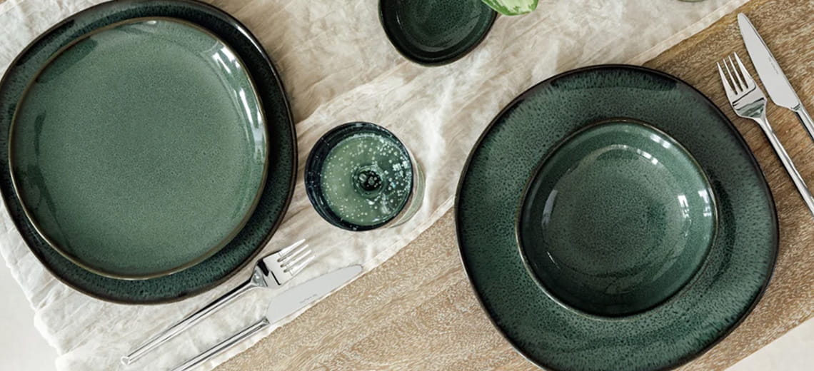 Apparecchiatura elegante con servizio di piatti in gres verde scuro, posate in acciaio e bicchieri coordinati su una tovaglia in lino chiara e tavolo in legno.