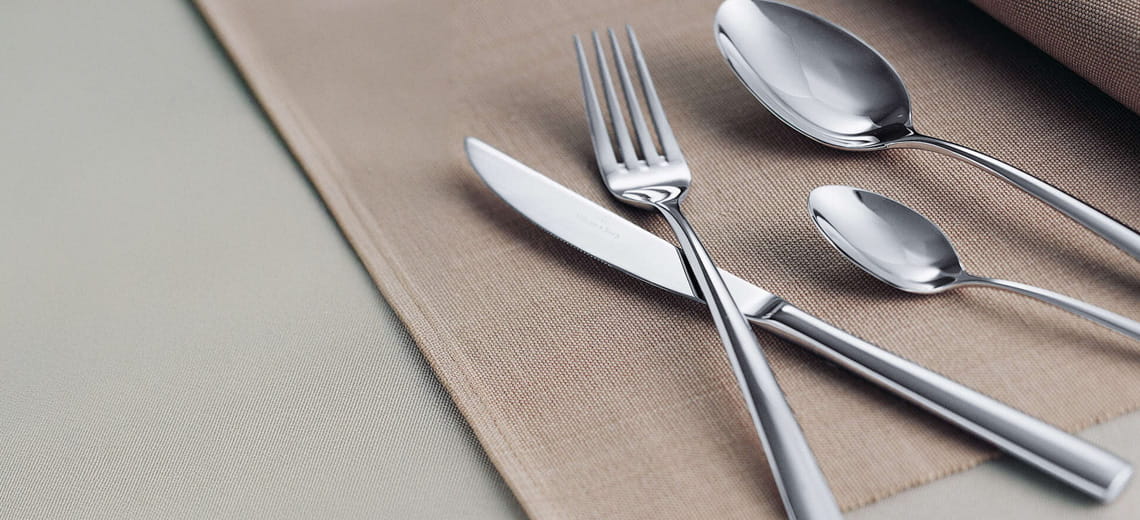 Set di posate in acciaio inox lucido composto da coltello, forchetta e cucchiai disposti su una tovaglietta di tessuto beige.